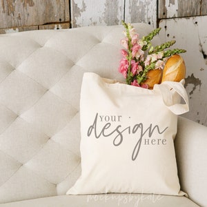 May include: A white canvas tote bag with the text "Your design here" printed on it. The bag is sitting on a white sofa with pink flowers and bread inside.