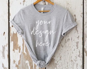 May include: A heather grey t-shirt with rolled up sleeves hanging on a wooden hanger against a whitewashed wood background. The shirt has the text "your design here" printed on the front in white.