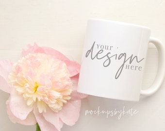 Könnte beinhalten: Eine weiße Keramiktasse mit Henkel, auf der der Text "Your Design Here" in grauer Schrift steht. Die Tasse steht auf einer weißen Oberfläche neben einer rosa Pfingstrose.