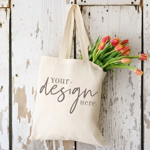 May include: A white canvas tote bag with the text "Your design here" hanging on a white wooden wall with a bouquet of red and yellow tulips.