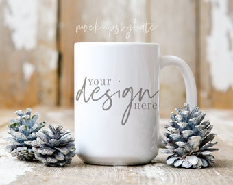 Puede incluir: Una taza de cerámica blanca con el texto "your design here" en escritura gris. La taza está flanqueada por dos piñas azul-grisáceas sobre una superficie de madera envejecida. El fondo es una pared de madera clara borrosa.