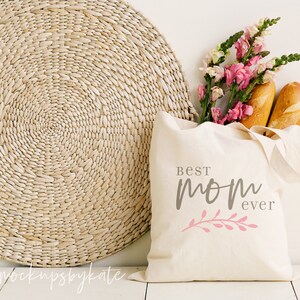 Op de afbeelding: Een witte canvas tote bag met de tekst "Best Mom Ever" in grijze en roze letters. De tas is gevuld met twee broden en een boeket roze en witte bloemen.