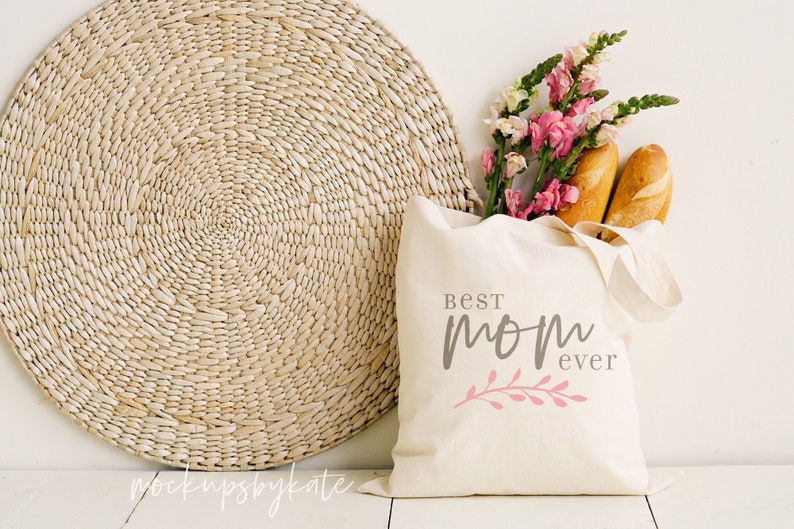 Op de afbeelding: Een witte canvas tote bag met de tekst "Best Mom Ever" in grijze en roze letters. De tas is gevuld met twee broden en een boeket roze en witte bloemen.
