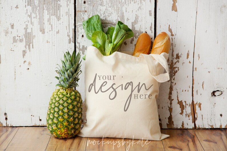 May include: A white canvas tote bag with the text "Your design here" on it. The bag is filled with groceries, including a head of lettuce, two baguettes, and a pineapple.