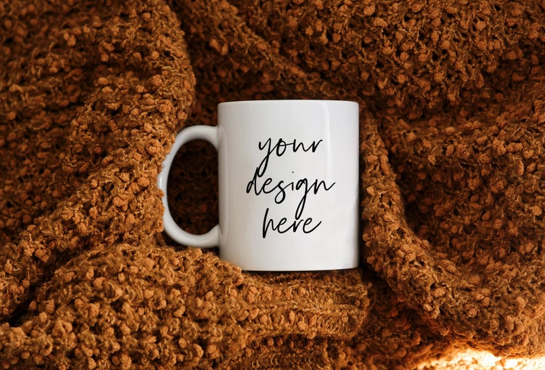 May include: A white ceramic mug with the text "your design here" in black script. The mug is set against a textured, brown, knitted blanket. The mug has a standard handle.