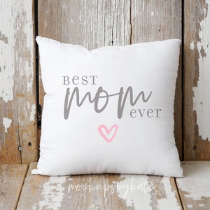 Puede incluir: Almohada blanca con el texto "Best Mom Ever" en letras grises y un coraz&oacute;n rosa. La almohada est&aacute; sobre una superficie de madera desgastada.