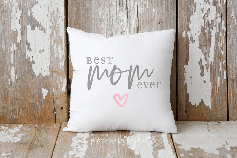 Puede incluir: Almohada blanca con el texto "Best Mom Ever" en letras grises y un coraz&oacute;n rosa. La almohada est&aacute; sobre una superficie de madera desgastada.