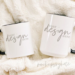 Może przedstawiać: Dwa białe ceramiczne kubki z czarnymi uchwytami i czarnymi obw&oacute;dkami. Kubki leżą na białym, puszystym kocu. Tekst "Your design here" jest napisany szarym kolorem na każdym kubku.