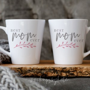 May include: Two white ceramic mugs with black lettering that reads "Best Mom Ever" and a pink floral design. The mugs are sitting on a wooden surface.