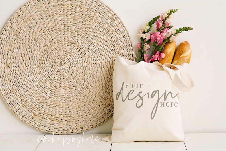 Op de afbeelding: Een witte canvas tote bag met de tekst "Your design here" in grijze schrijfletters. De tas is gevuld met twee broden en een boeket roze en witte bloemen. De tas ligt op een wit oppervlak naast een ronde geweven mat.