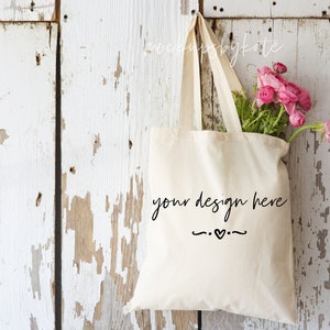 May include: A cream-colored tote bag with the text "your design here" in a handwritten font, hanging against a distressed white wooden background. The bag is filled with pink flowers and green foliage.