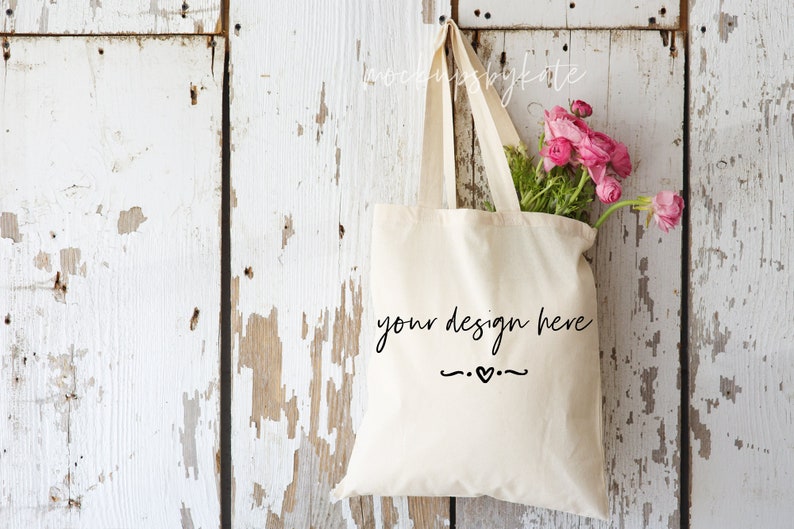 May include: A cream-colored tote bag with the text "your design here" in a handwritten font, hanging against a distressed white wooden background. The bag is filled with pink flowers and green foliage.