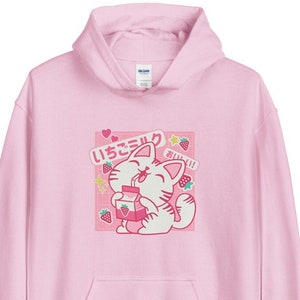 K&ouml;nnte beinhalten: Hellrosa Kapuzenpullover mit einer Grafik einer Cartoon-Katze, die Erdbeermilch trinkt. Die Grafik enth&auml;lt japanischen Text, Erdbeeren und Sterne. Der Hoodie hat eine Vordertasche und eine Kapuze.