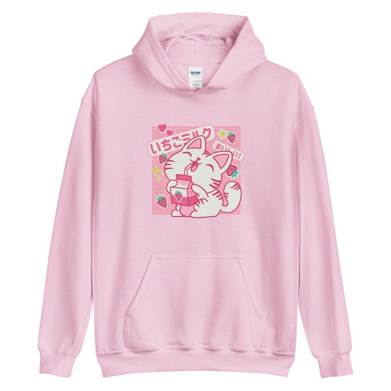 K&ouml;nnte beinhalten: Hellrosa Kapuzenpullover mit einer Grafik einer Cartoon-Katze, die Erdbeermilch trinkt. Die Grafik enth&auml;lt japanischen Text, Erdbeeren und Sterne. Der Hoodie hat eine Vordertasche und eine Kapuze.