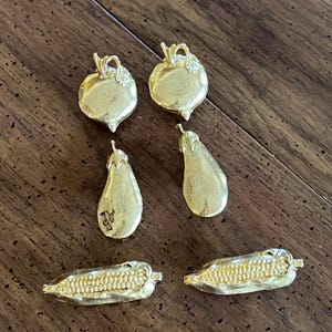May include: A collection of six gold-toned metal charms shaped like vegetables. The set includes two round turnip-shaped charms, two elongated eggplant-shaped charms, and two corn-on-the-cob charms, all featuring textured surfaces and metallic finishes.