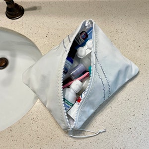 May include: A white zippered toiletry bag with blue stitching is open and filled with various toiletries, including a toothbrush, toothpaste, mouthwash, and hand sanitizer.