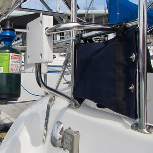 May include: A white metal bracket mounted on a white boat deck with a stainless steel railing and a blue canvas bag hanging from it. A green propane tank is attached to the railing.