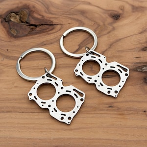 May include: Two silver-colored keychains featuring a detailed engine part design. Each keychain has a circular key ring attached to a metal pendant with multiple holes and cutouts. The keychains are displayed on a wooden surface.