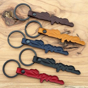 May include: Five leather keychains featuring the word Supra embossed in a script font. Each keychain has a black metal split ring attached by a dark metal rivet. The leather colors include dark brown, tan, navy blue, black, and red.