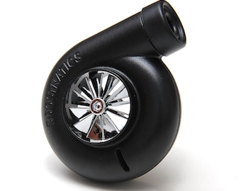 Puede incluir: Un modelo de turbocompresor negro mate con una rueda de turbina central de acabado cromado y múltiples aspas. La carcasa tiene grabado el texto BOOSTNATICS e incluye un puerto de salida cilíndrico. La palabra TURBO aparece impresa debajo.