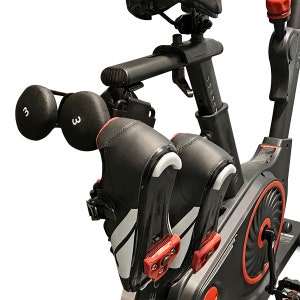 Puede incluir: Una bicicleta de ejercicio negra con detalles rojos. Dos zapatillas de ciclismo negras con detalles blancos están sujetas a los pedales. Dos pesas negras con el número "3" impreso en ellas están sujetas al manillar.