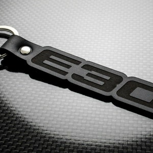 Puede incluir: Llavero de cuero negro con un llavero plateado. El llavero presenta las letras "E30" en relieve en forma rectangular. El diseño es simple y moderno, adecuado para los entusiastas de los coches.