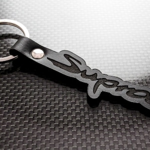 May include: Black leather keychain featuring the word "Supra" in a stylized font. It has a silver metal keyring and a small silver rivet. The background is a textured, dark grey surface.