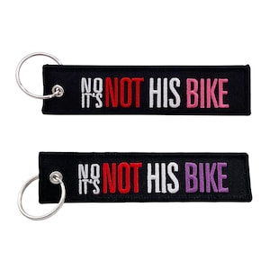 May include: Black fabric keychain with a silver key ring. The keychain has white text that says "It's Not His Bike" with the word "Not" in red and "Bike" in pink.
