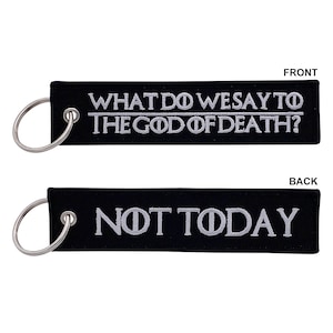 May include: Black keychain with white embroidery. The front reads "What do we say to the god of death?" and the back reads "Not today."