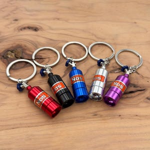 Puede incluir: Colección de cinco llaveros de botellas NOS en miniatura en rojo, negro, azul, plateado y morado. Cada llavero presenta un llavero plateado, una válvula azul y la palabra "NOS" en naranja. Se muestran sobre una superficie de madera.
