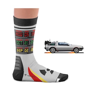 May include: A pair of gray and black socks with a design of a time machine from the movie Back to the Future. The socks feature a digital display with the date "NOV 05 1955", "OCT 21 2015", and "SEP 02 1885". The socks also feature a graphic of the time machine with a red, yellow, and black design.