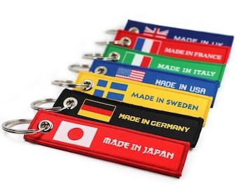 Pode incluir: Um conjunto de sete chaveiros com bandeiras e o texto "Made in" seguido do país de origem. Os chaveiros são vermelho, azul, amarelo, verde, preto, branco e azul claro.