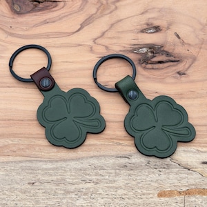 Peut inclure: Deux porte-clés en cuir vert en forme de trèfle à trois feuilles. Chaque porte-clés a un anneau en métal noir et une languette en cuir marron ou vert. Le motif de trèfle est en relief sur le cuir.