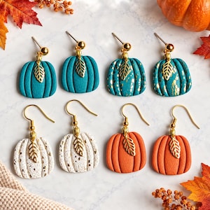 May include: Four pairs of pumpkin-shaped earrings featuring gold-tone metal leaf charms. The set includes solid teal, gold-flecked teal, gold-flecked white, and solid orange pumpkins. Each earring is suspended from a gold-tone metal hook or post.