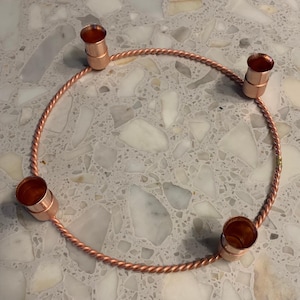 Peut inclure: Une couronne de l'Avent de couleur cuivre avec quatre bougeoirs. La couronne est une conception de corde torsadée, et les bougeoirs sont cylindriques. La couronne est conçue pour contenir quatre bougies pour la saison de l'Avent.