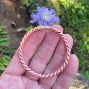 May include: A circular bangle bracelet crafted from a single continuous strand of twisted copper wire. The metal has a smooth, metallic pinkish-orange finish with a small, slightly textured gold-colored solder joint visible at the top of the loop.