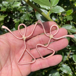 May include: Two copper-colored wire figures, resembling dancing people, are held in a hand against a backdrop of green foliage. The figures have looped heads, outstretched arms, and triangular or curved lower bodies. The figures are connected at the top with a gold-colored joint.