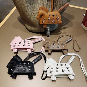 May include: A collection of five horse-shaped leather bag charms, each featuring silver-toned metal studs and a matching leather strap. The charms are available in tan, light pink, brown, black, and white, with one attached to a textured leather bag.