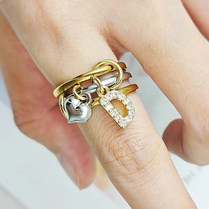 May include: A multi-band ring featuring a combination of gold, silver, and rose-gold tones. The ring is adorned with two dangling charms: a smooth, polished silver-tone heart and a letter D charm encrusted with small, clear, sparkling stones.