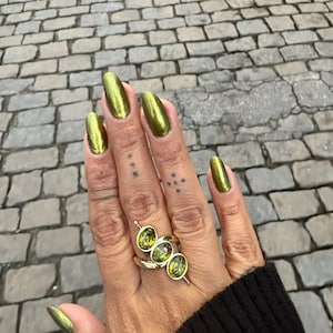 May include: A gold-toned ring featuring three oval-shaped green gemstones arranged in a diagonal row. The ring is worn on a finger with metallic olive green nail polish. The band is thin and polished, showcasing a simple, elegant design.