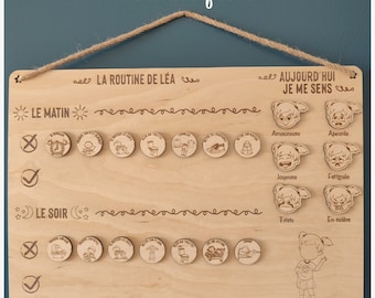 May include: A wooden board with a routine chart for a child. The chart includes a section for morning and evening routines, with icons for activities like getting dressed, brushing teeth, and going to bed. There is also a section for the child to choose how they are feeling, with icons for different emotions like happy, sad, and angry. The text on the board reads "La Routine de Lea" and "Aujourd'hui Je Me Sens".