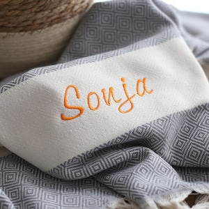 Puede incluir: Una manta tejida gris y blanca con un patr&oacute;n de diamantes y flecos. La manta tiene una franja blanca con el nombre "Sonja" bordado en hilo naranja. Una cesta tejida est&aacute; en el fondo.