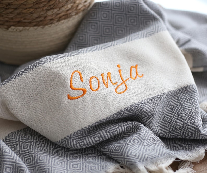 Puede incluir: Una manta tejida gris y blanca con un patr&oacute;n de diamantes y flecos. La manta tiene una franja blanca con el nombre "Sonja" bordado en hilo naranja. Una cesta tejida est&aacute; en el fondo.