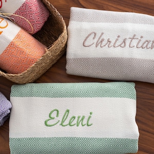 Puede incluir: Toallas de playa personalizadas con nombres bordados. Las toallas tienen un panel central blanco, bordes con dise&ntilde;o de diamantes y rayas de colores. Una toalla est&aacute; en una cesta. Los nombres bordados son Kylie, Christiana y Eleni.