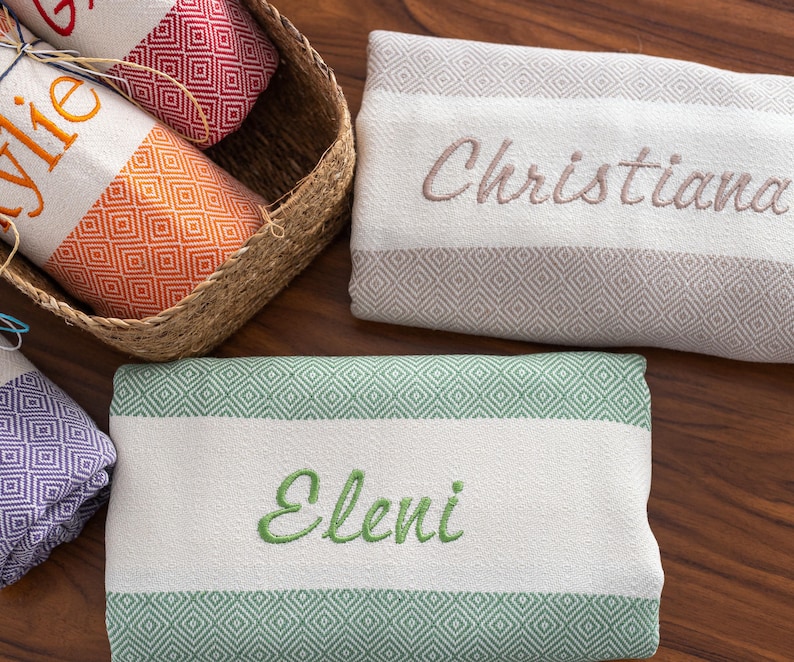 Puede incluir: Toallas de playa personalizadas con nombres bordados. Las toallas tienen un panel central blanco, bordes con dise&ntilde;o de diamantes y rayas de colores. Una toalla est&aacute; en una cesta. Los nombres bordados son Kylie, Christiana y Eleni.