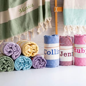 Puede incluir: Toallas turcas enrolladas en varios colores, incluyendo verde, amarillo, azul y morado. Algunas toallas están personalizadas con nombres como "Monica", "Collin", "Jenn" y "Ruby". Las toallas tienen un patrón de diamantes y flecos.