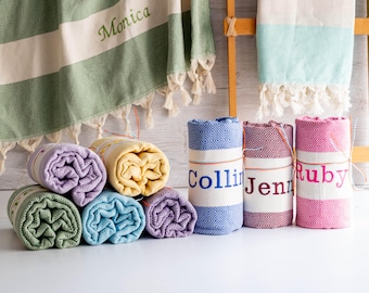 May include: Rolled Turkish towels in various colors, including green, yellow, blue, and purple. Some towels are personalized with names like "Monica," "Collin," "Jenn," and "Ruby." The towels have a diamond pattern and fringe.
