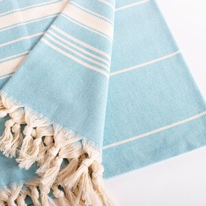 Puede incluir: Una toalla doblada a rayas azul claro y blancas con flecos color crema. La toalla tiene un tejido texturizado y est&aacute; hecha de un material suave. Las rayas son horizontales. Los flecos est&aacute;n anudados.