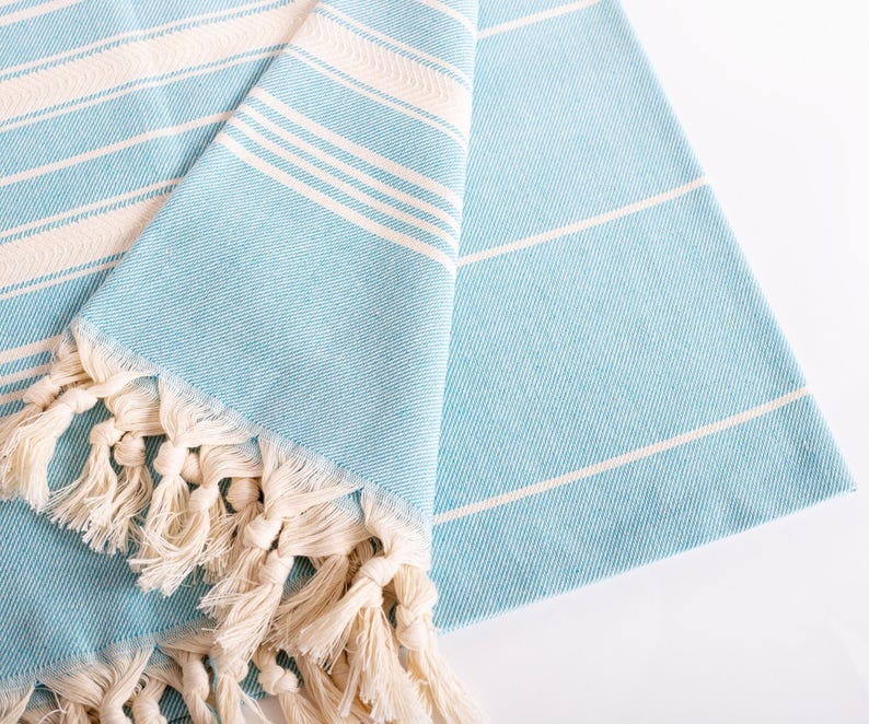 Puede incluir: Una toalla doblada a rayas azul claro y blancas con flecos color crema. La toalla tiene un tejido texturizado y est&aacute; hecha de un material suave. Las rayas son horizontales. Los flecos est&aacute;n anudados.