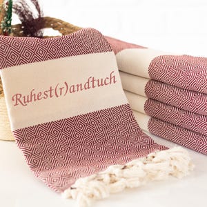Puede incluir: Toallas burdeos y crema plegadas con un patr&oacute;n geom&eacute;trico de diamantes. Una toalla est&aacute; drapeada hacia adelante, mostrando el texto "Ruhest(r)andtuch" en burdeos. Las toallas tienen flecos color crema.